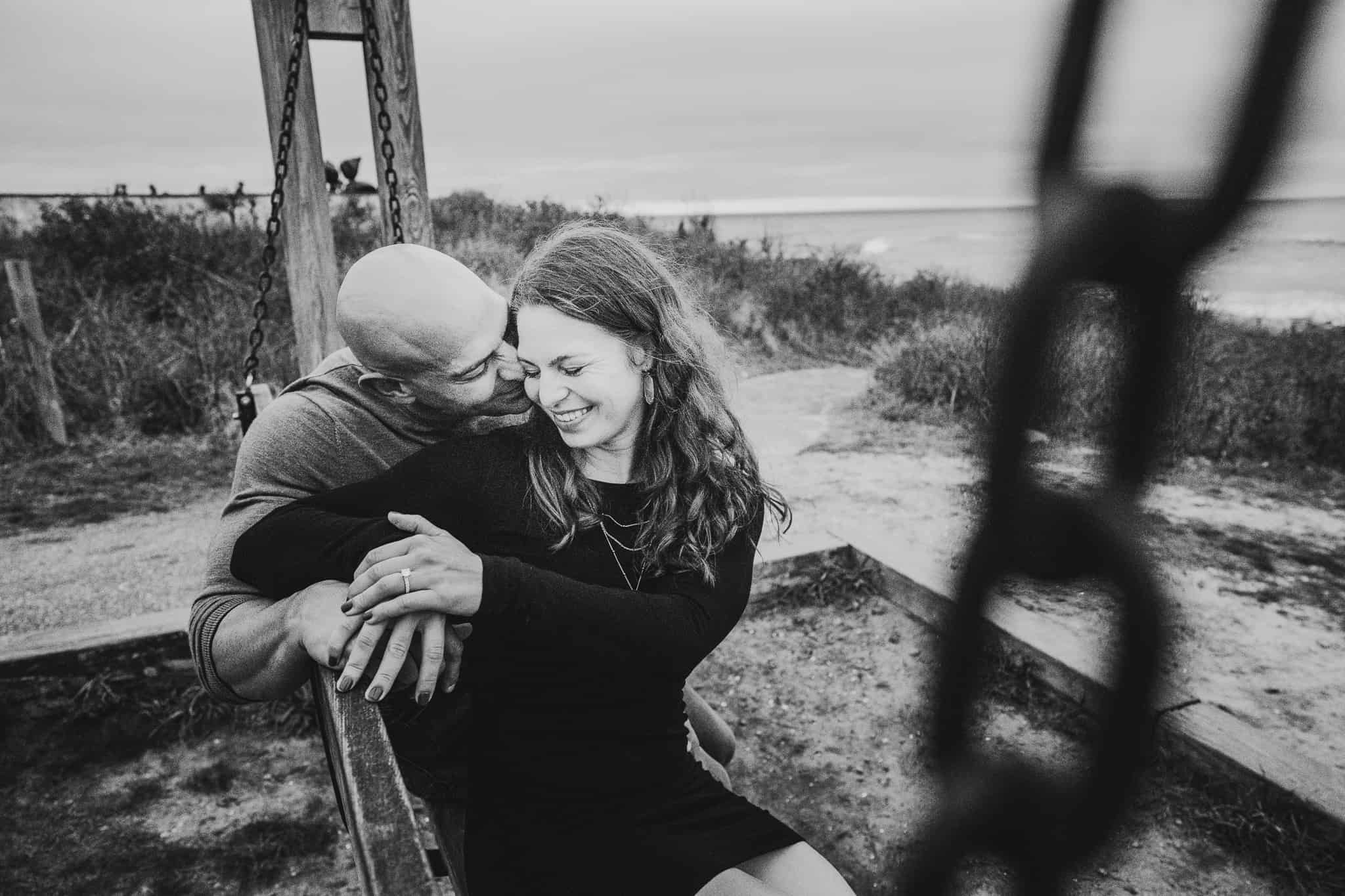 Couple cuddling and laughing together on a swing bench at the Montauk lighthouse beach in Long Island New York during their engagement and proposal session