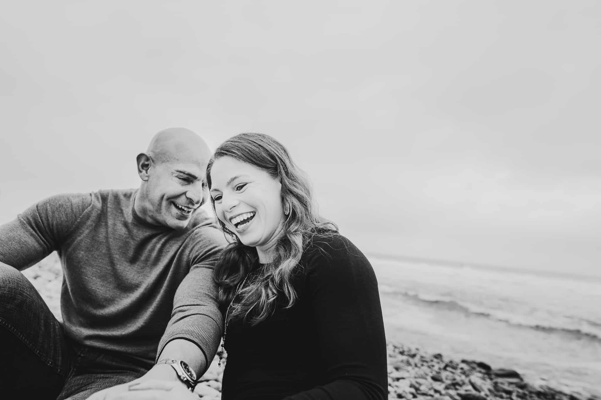 Black and white photo of a couple laughing and giggling together during their engagement session on the beach with the water in the background at Montauk lighthouse in Long Island New York