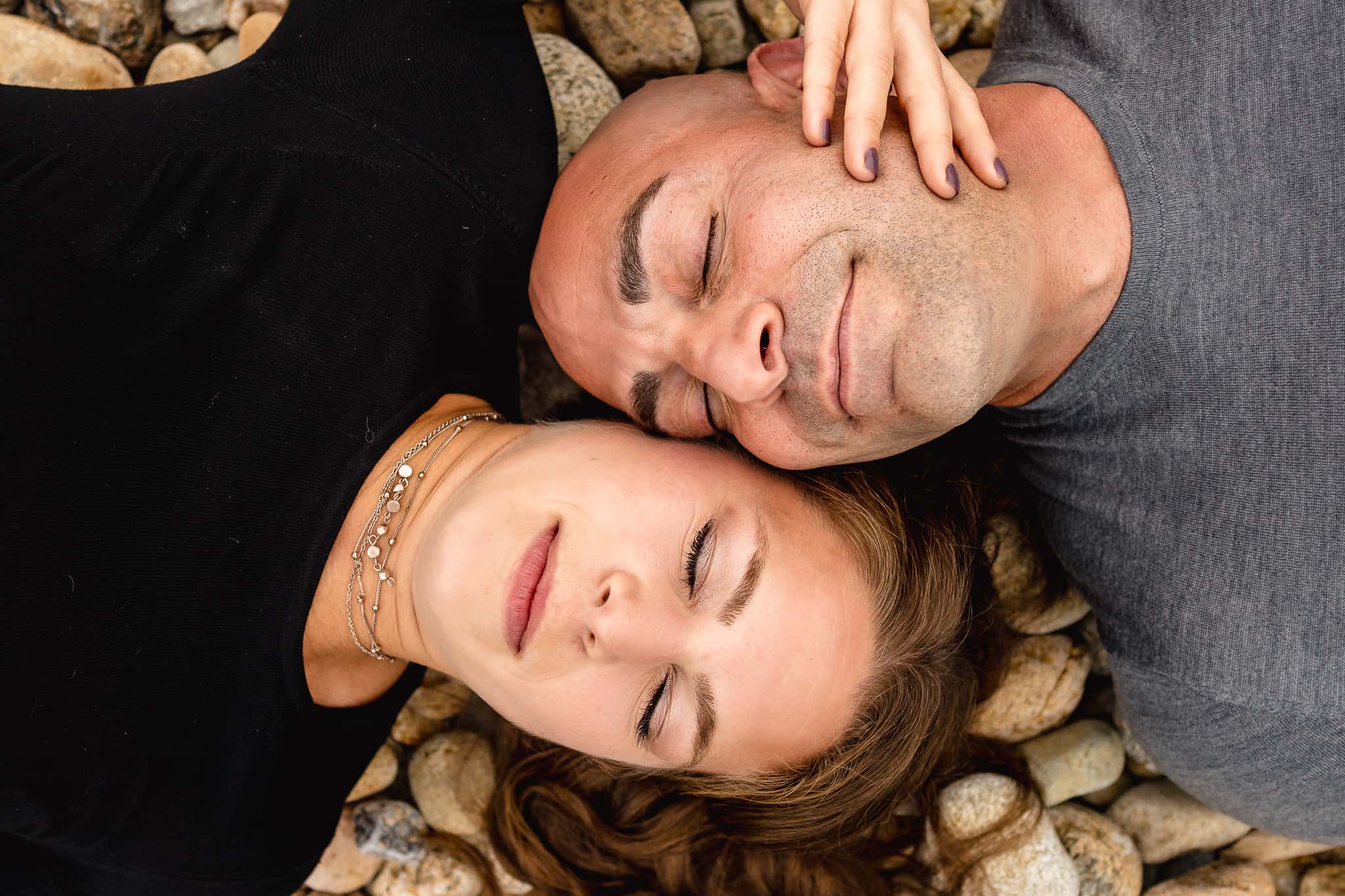 Couple laying down on the beach at Montauk lighthouse resting their heads together with their eyes closed peacefully next to Long Island New York during their engagement session