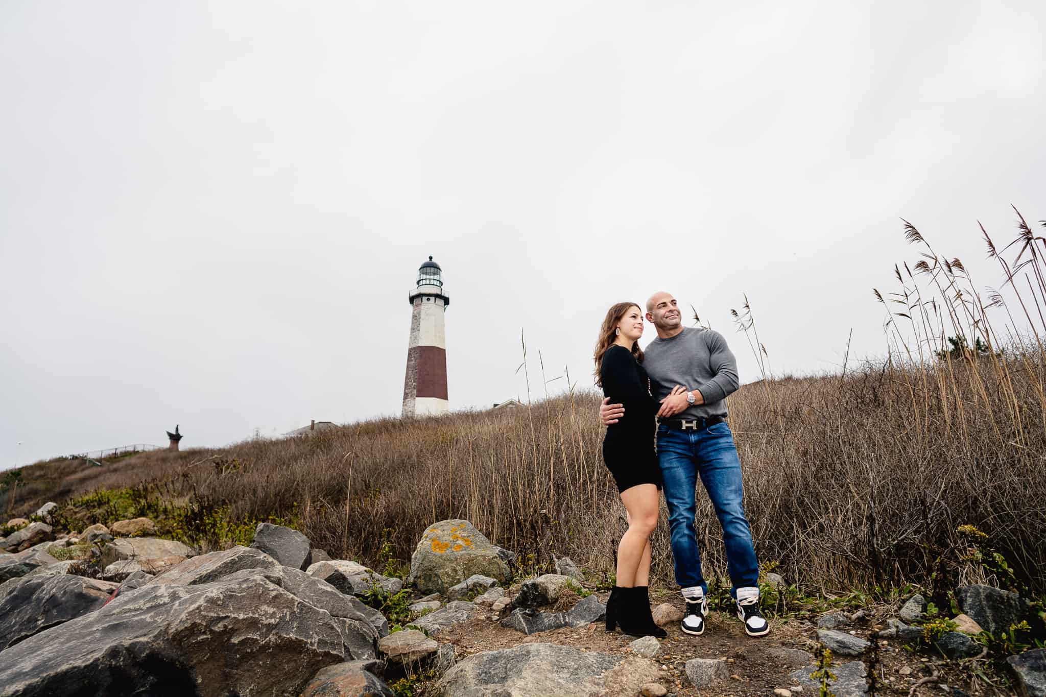 Wide landscape portrait of a couple during their engagement session holding hands and cuddling up in front of the Montauk lighthouse with a Gray sky in Long Island New York