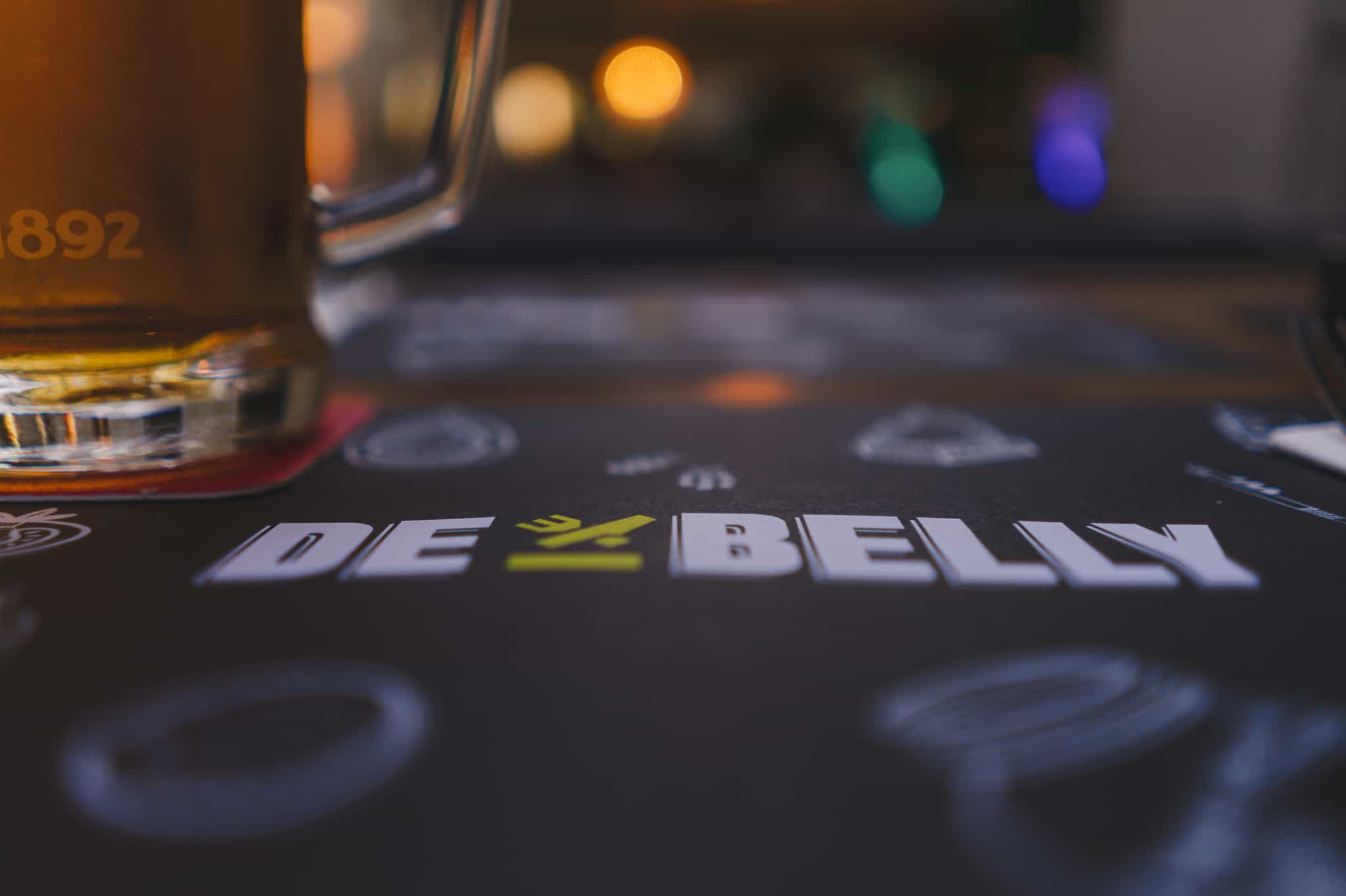 The logo of the restaurant De Belly in split Croatia with a beer and light bokeh in the background