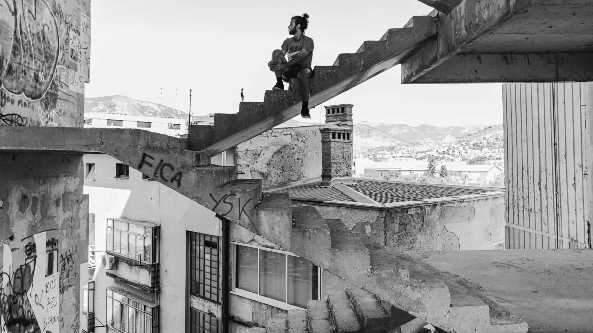 A hipster sitting at the top of a staircase with no walls in the Mostar sniper tower with Bosnia and Herzegovina in the background