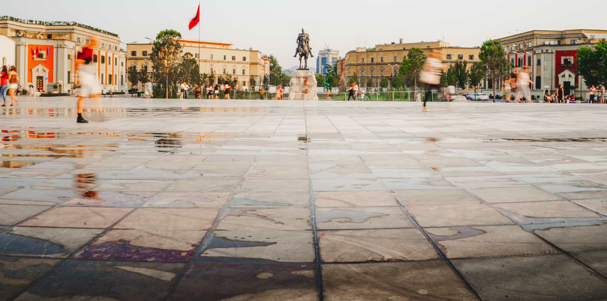 Wide landscape shot of skanderbeg square in Tirana Albania shot from low with water glistening on the ground and all of the people in motion blur