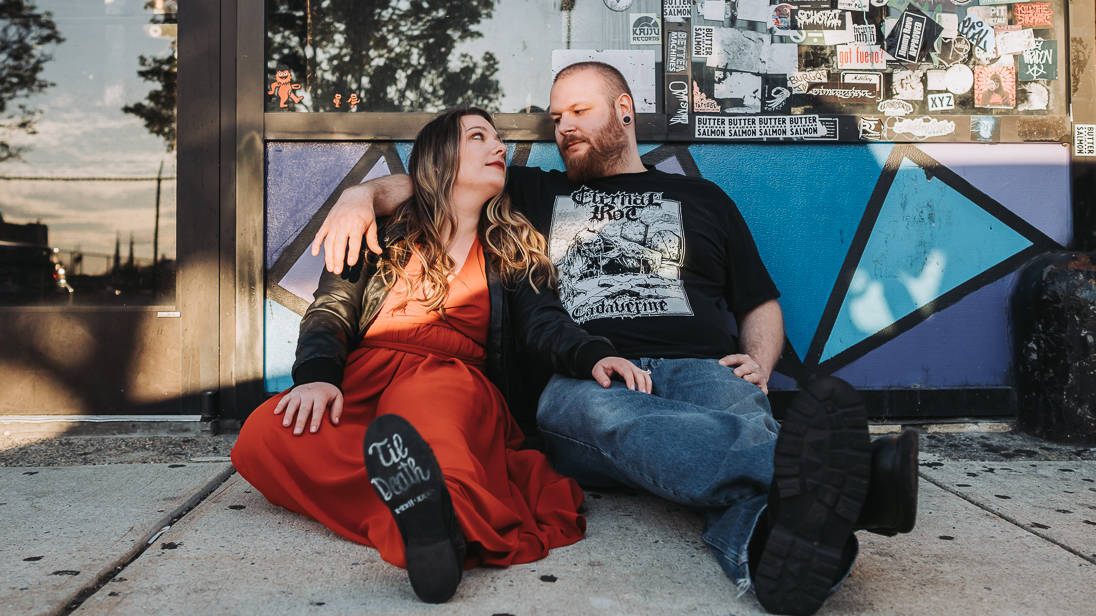 Engaged couple sitting and posed on the ground in front of a music store in fishtown Philadelphia looking lovingly at each other during sunset