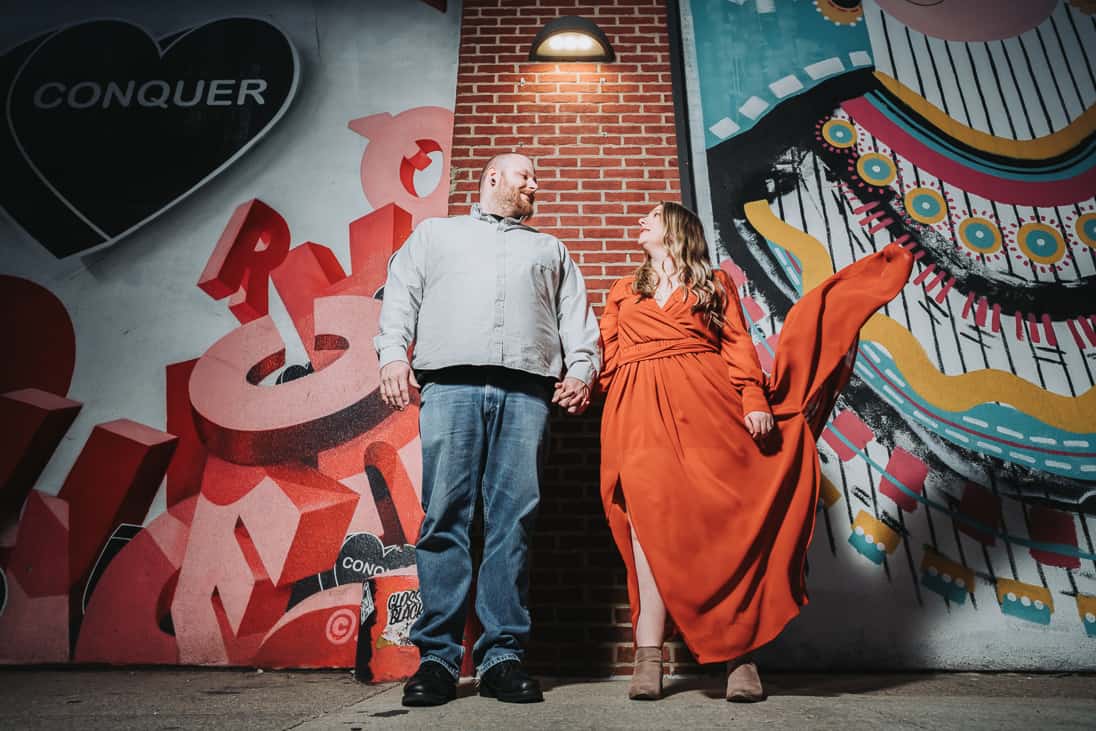 Engagement photo of a couple standing in front of graffiti and fishtown Philadelphia at night time dramatically lit with her dress flying in the air