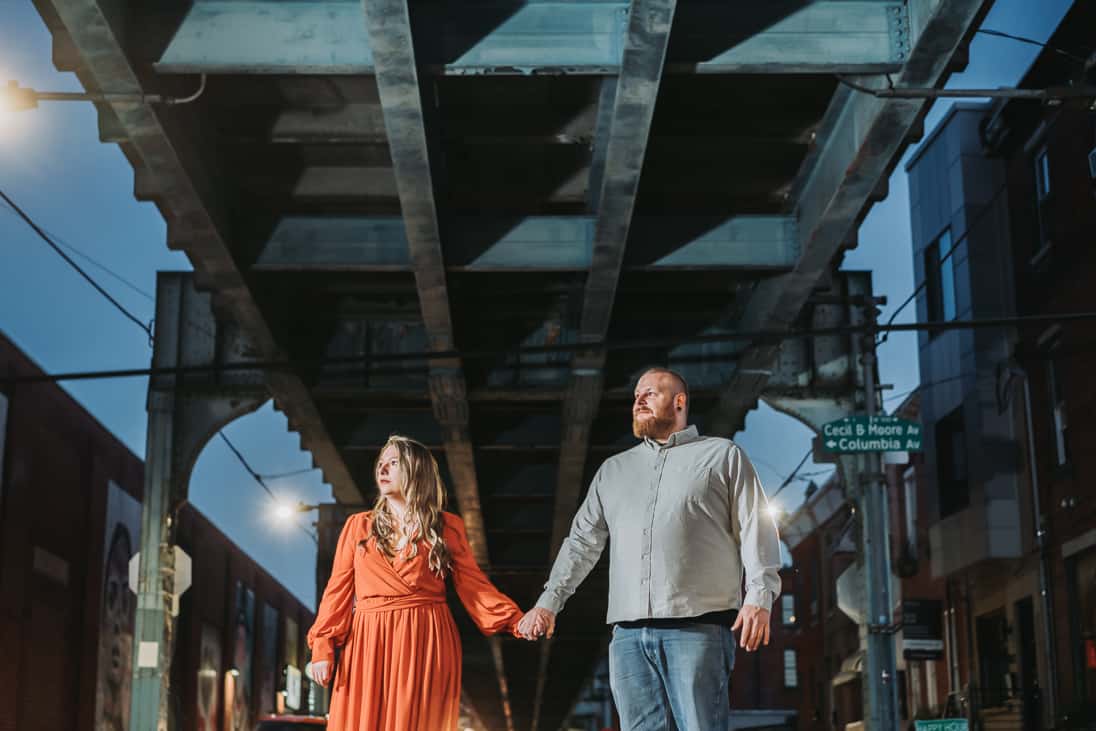 Dramatic engagement photo of a couple standing under the SEPTA in fishtown Philadelphia looking towards the sky