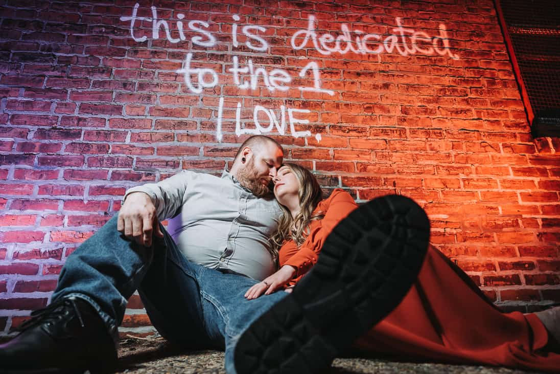 Dramatically lit purple and red photo of a engaged couple posed and sitting on the ground in front of graffiti in fishtown Philadelphia that reads "This s dedicated to the one I love."