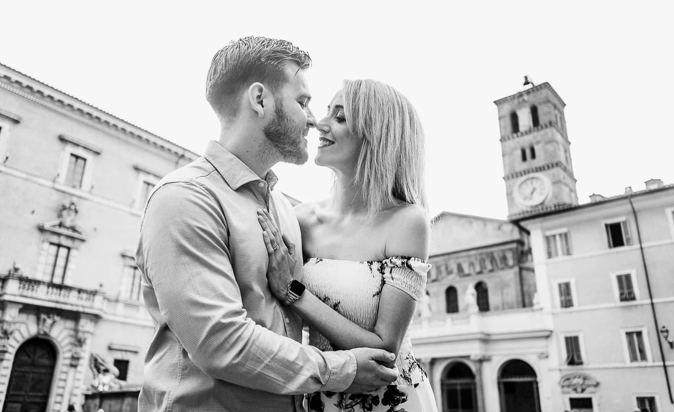 Black and white photo of a couple nestling noses with piazza de Santa Maria in the background during an engagement photo shoot in Rome Italy